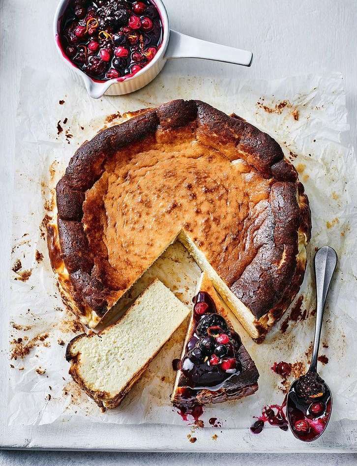 Overhead shot of a Basque cheesecake with a slice cut, topped with berry sauce, and a spoonful of sauce on a white background.