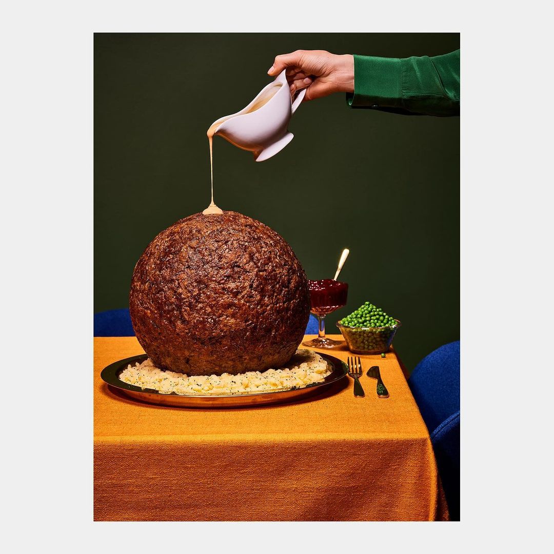 A giant meatball on a platter is drizzled with sauce from a gravy boat held by a green-sleeved arm, next to peas and cranberry sauce.