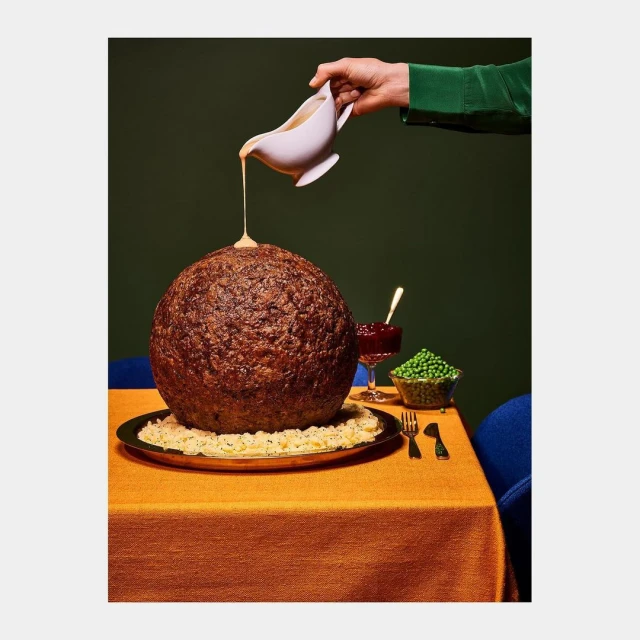 A giant meatball on a platter is drizzled with sauce from a gravy boat held by a green-sleeved arm, next to peas and cranberry sauce.