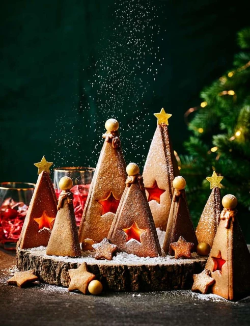Festive arrangement of gingerbread Christmas trees with star cutouts on a wooden base, dusted with powdered sugar, evergreen boughs in background.