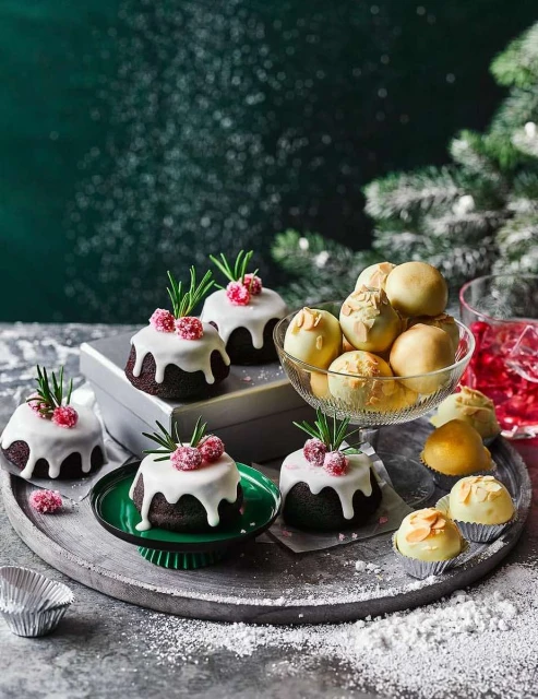 Festive dessert display with mini chocolate cakes, cream-filled bites, and a glass of red liquid on a silver tray with snowy backdrop.