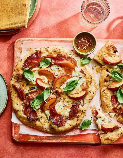 Overhead shot of peach and prosciutto pizza on a wooden board, garnished with basil, alongside a small bowl of sauce and a glass of sparkling water.