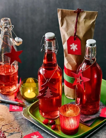 Holiday-themed still life features three glass bottles filled with a red liquid on a green tray, along with gift bag and candles.
