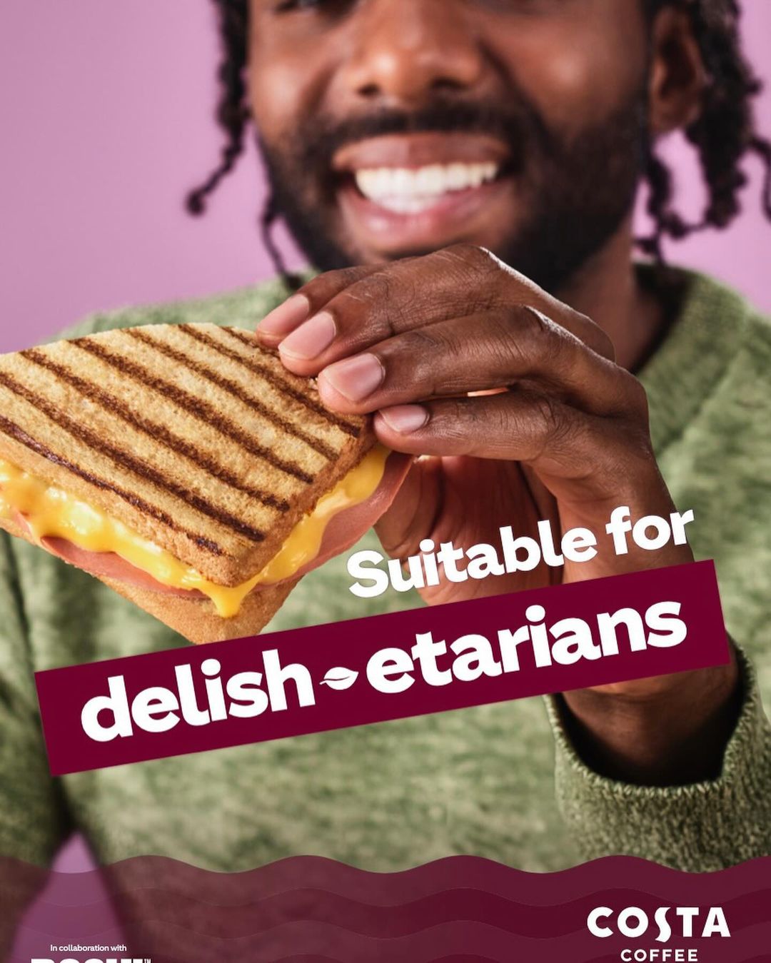 A smiling Black male holds a toasted sandwich in front of a pink background for a Costa Coffee ad promoting vegetarian options.