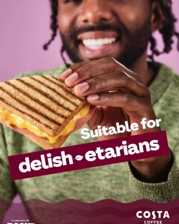 A smiling Black male holds a toasted sandwich in front of a pink background for a Costa Coffee ad promoting vegetarian options.