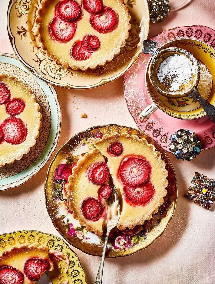 Overhead shot of several strawberry tarts on antique plates. The tarts have a creamy base and sliced strawberries on top, presented with vintage tableware.