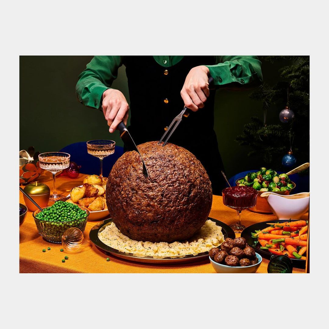 Person cuts into a large spherical meat dish at a festive table with various food bowls, glasses, and a Christmas tree in the background.