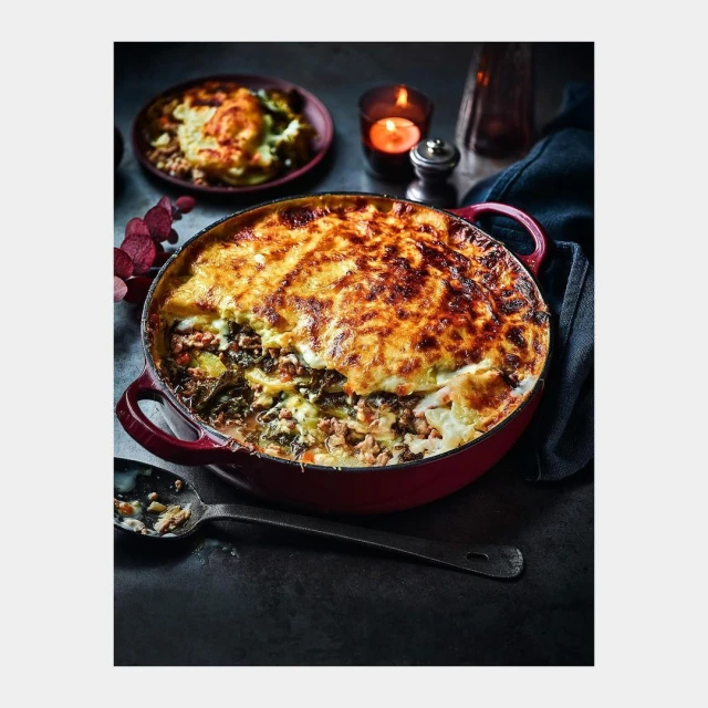 Overhead shot of a rustic red cast-iron pot filled with homemade moussaka, garnished with herbs, alongside candle and stoneware dish.