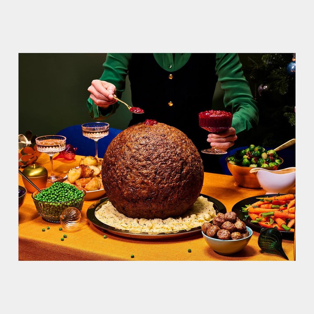 Person adding cranberry sauce to a giant meatball centerpiece on a holiday table with roasted vegetables and crystal glasses.