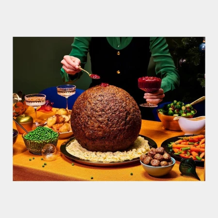 Person adding cranberry sauce to a giant meatball centerpiece on a holiday table with roasted vegetables and crystal glasses.
