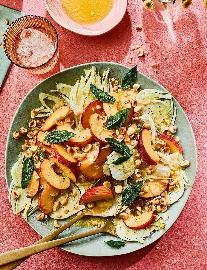 Overhead shot of a peach and fennel salad on a green plate with hazelnuts and sage, beside a glass of water and a bowl of dressing.