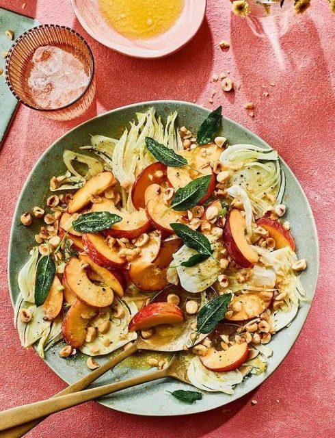 Overhead shot of a peach and fennel salad on a green plate with hazelnuts and sage, beside a glass of water and a bowl of dressing.