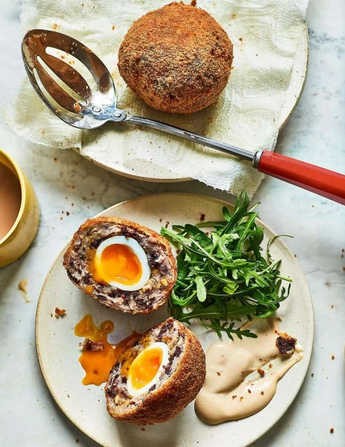 Overhead close-up of sliced Scotch egg with runny yolk, whole Scotch egg, greens, sauce, and a spoon on a speckled surface.