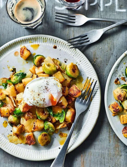 Overhead shot of two plates with fried potatoes, brussel sprouts and poached egg, garnished with sauce, served on rustic wooden table with forks.