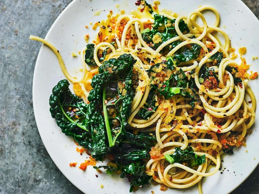 Overhead shot showcases a plate of spaghetti with kale, breadcrumbs, and a glossy sauce against a textured grey backdrop, creating a culinary composition.