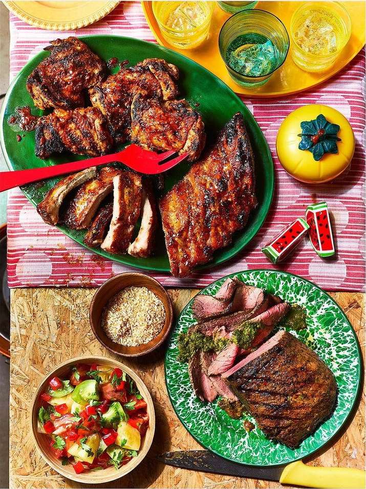 Overhead shot of grilled meat and sides on colorful plates and placemats, including ribs, chicken, steak, and a vibrant tomato salad.