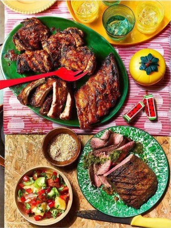 Overhead shot of grilled meat and sides on colorful plates and placemats, including ribs, chicken, steak, and a vibrant tomato salad.