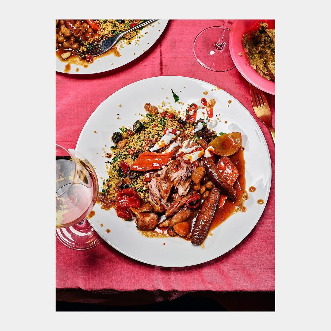 Overhead shot of a white plate with sausages, couscous, meat, peppers, and other foods, with a glass of wine on a pink tablecloth.