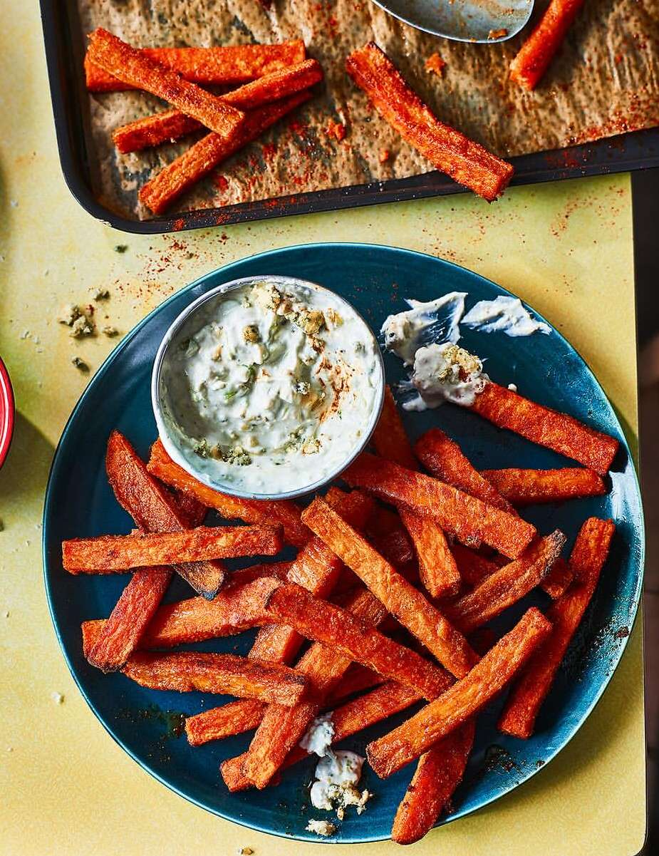 Overhead shot of sweet potato fries served with dipping sauce on a blue plate and baking sheet with parchment paper.