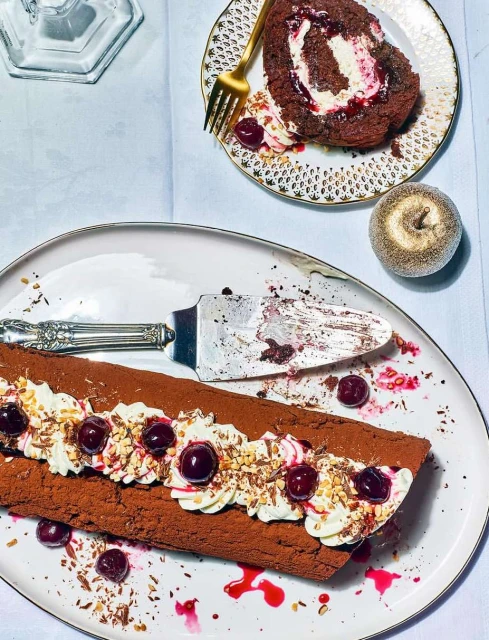 Overhead shot of black forest cake roll on an ornate plate with cherries, whipped cream, and chocolate shavings. Cake server included.
