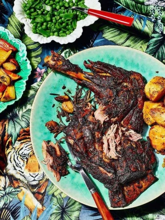 Overhead shot of roast lamb leg on a turquoise plate, accompanied by roasted potatoes and green beans, on a patterned tablecloth.