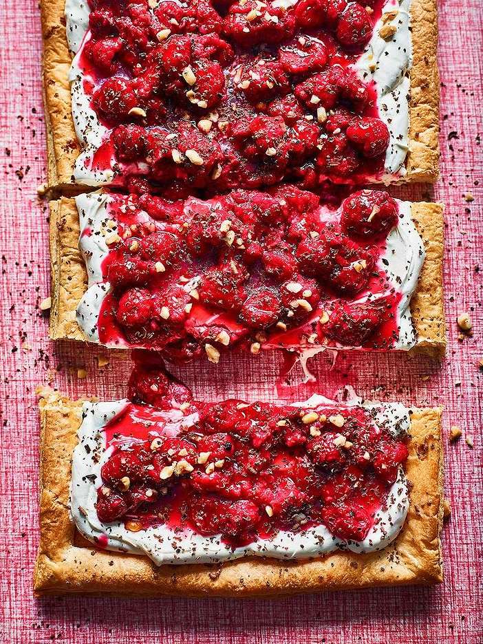 Overhead shot of rectangular puff pastry tarts with white cream, raspberries, and chopped nuts on a pink textured surface.