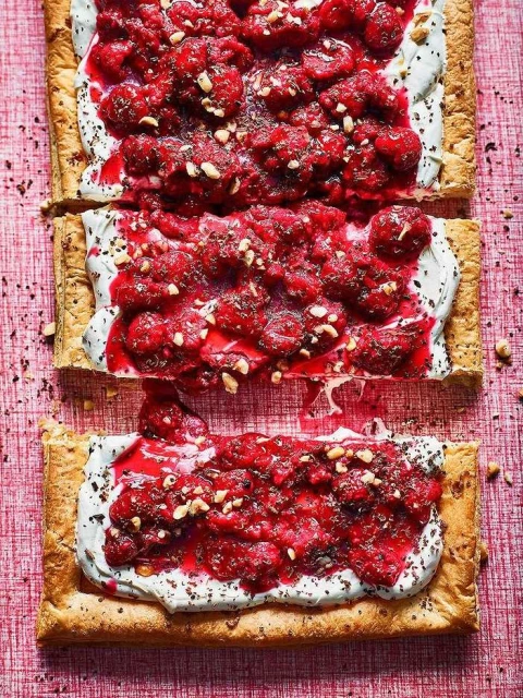 Overhead shot of rectangular puff pastry tarts with white cream, raspberries, and chopped nuts on a pink textured surface.