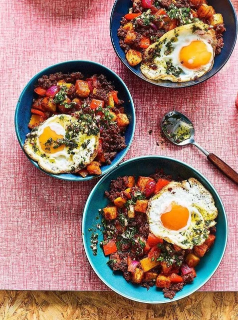 Overhead shot of three bowls of food with potatoes, meat, peppers, and fried eggs, plated on a pink tablecloth, studio lighting.