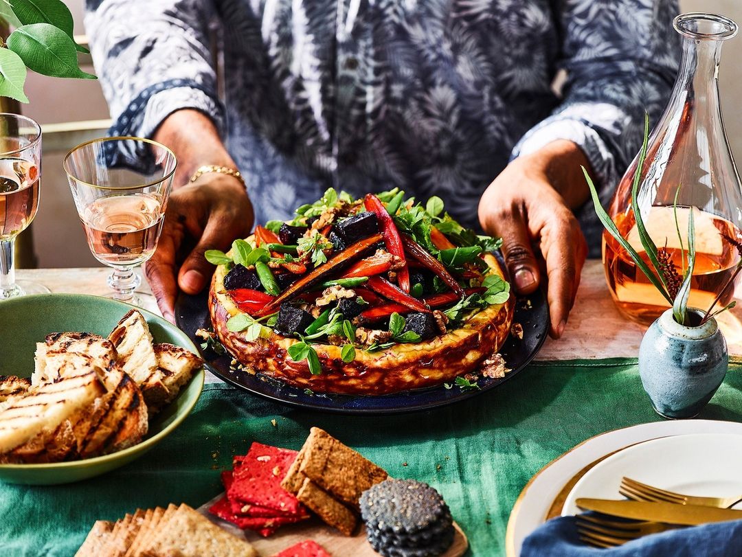 Overhead food photography of a person serving a savory cheesecake topped with greens, peppers, and beets, surrounded by appetizers.