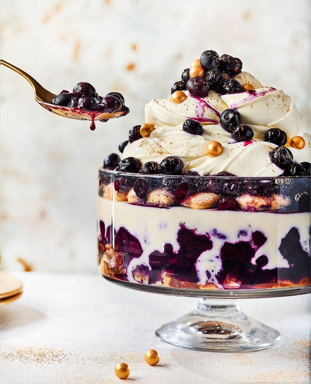 Close-up of a trifle dessert on a footed bowl with layers of sponge cake, cream, blueberries, and gold sprinkles being served with a golden spoon.