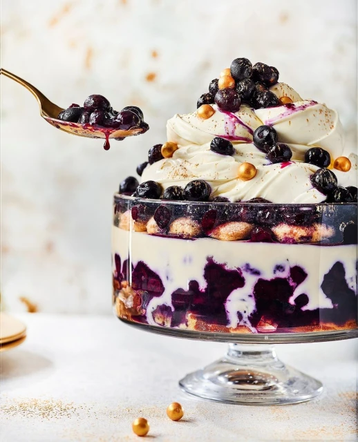 Close-up of a trifle dessert on a footed bowl with layers of sponge cake, cream, blueberries, and gold sprinkles being served with a golden spoon.