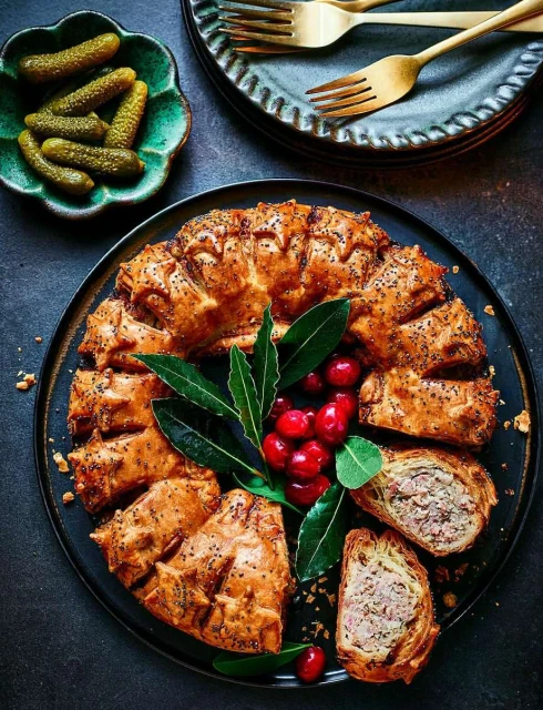 Overhead shot of a savory meat pie garnished with cranberries and bay leaves, along with a bowl of pickles and stacked plates.