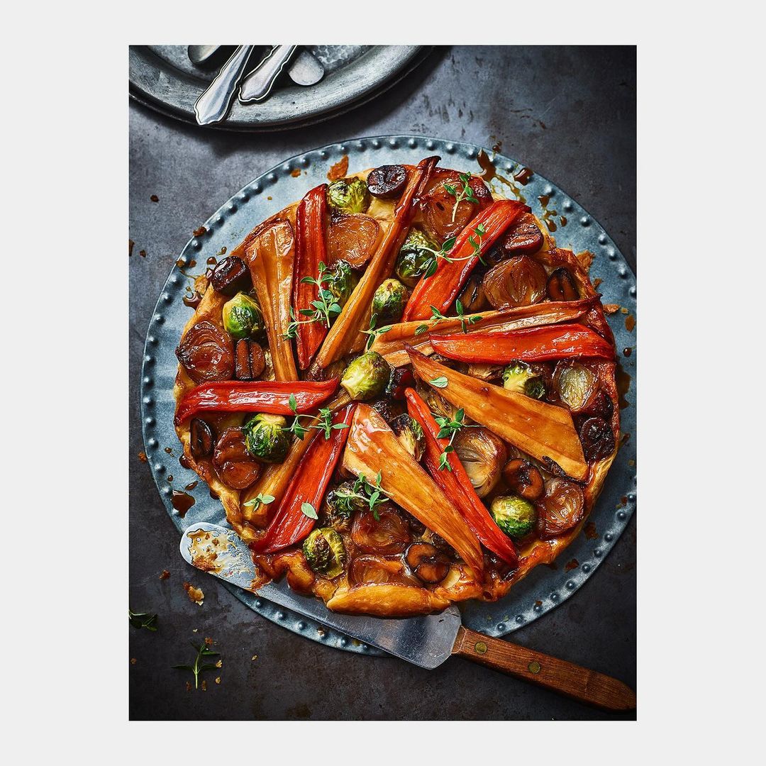 Overhead shot of a glazed vegetable tarte tatin featuring root vegetables and brussel sprouts on a rustic metal platter with serving utensils.