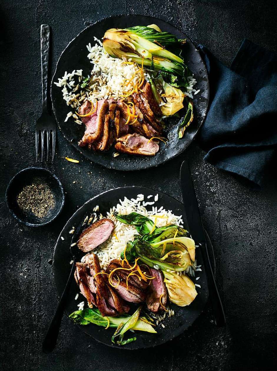 Two plates of Asian-inspired cuisine featuring seared duck breast, rice, and bok choy, presented on a dark, textured surface with utensils.