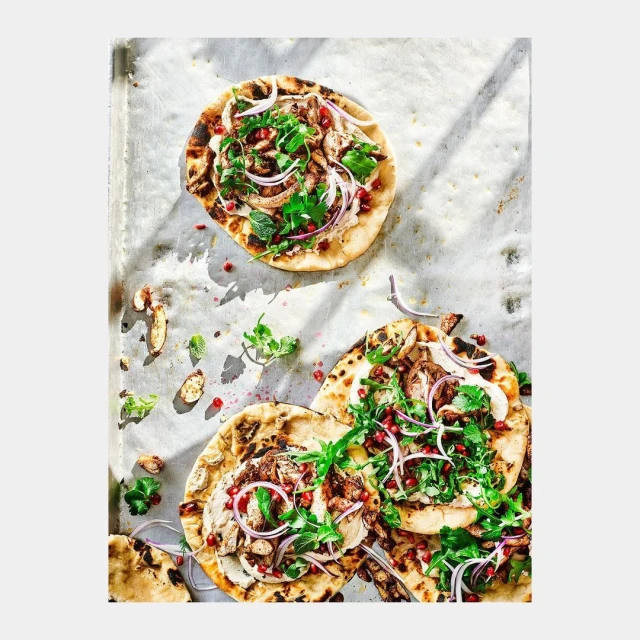 Overhead shot of three flatbreads topped with meat, onions, pomegranate seeds, and parsley on a silver baking sheet with sunlight.