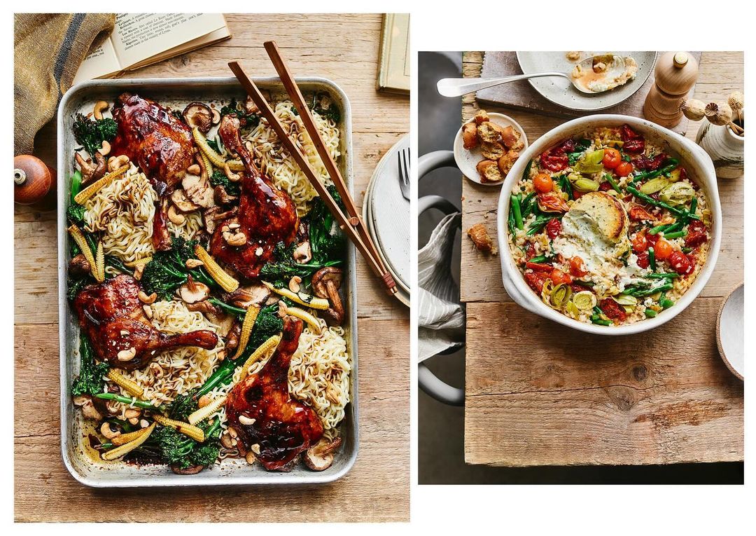 Overhead shot of glazed duck legs with noodles and vegetables on a metal tray, plus a casserole dish of vegetables with a spoon on a wood surface.