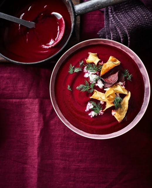 Overhead shot of beet soup in a pink bowl with beet chips and dill, next to a pot of soup with a ladle on a burgundy tablecloth.