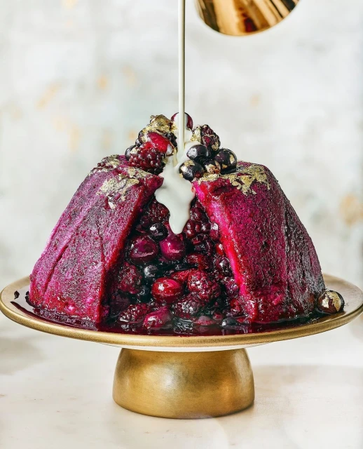 Festive berry pudding on a gold cake stand is drizzled with white sauce, creating a visually appealing and delectable food image.