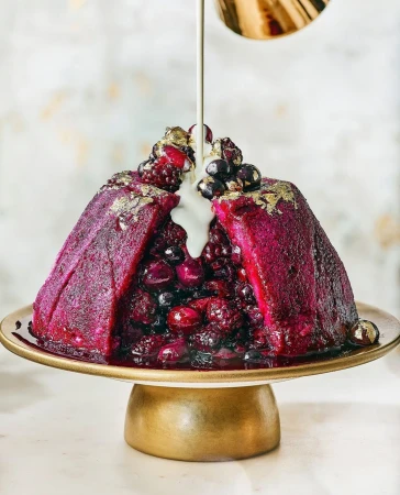 Festive berry pudding on a gold cake stand is drizzled with white sauce, creating a visually appealing and delectable food image.