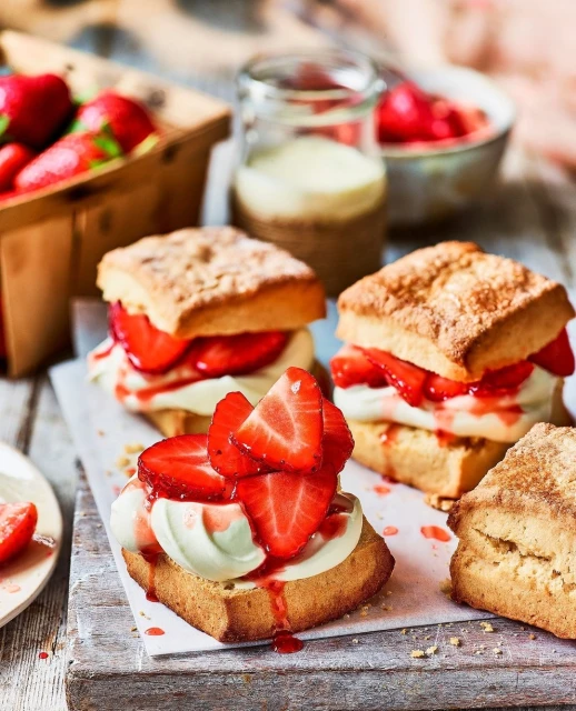 Three strawberry shortcakes on a white parchment paper on a wooden surface, with fresh strawberries and cream in the background.