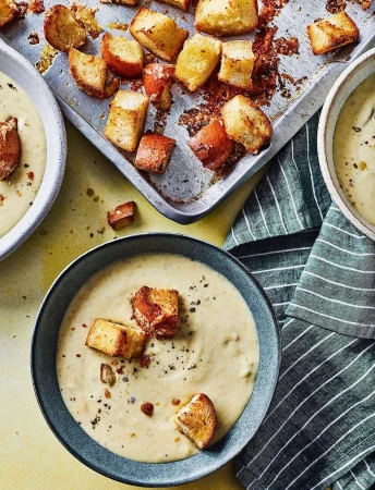 Overhead shot of bowls of soup with croutons and a metal sheet pan of more croutons, alongside a striped kitchen towel.