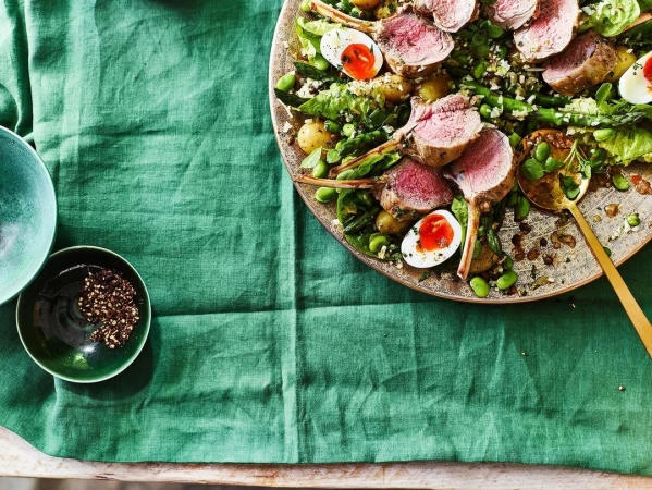 Overhead shot of a plate with sliced lamb chops, boiled eggs, green beans, and couscous salad on a green linen tablecloth.
