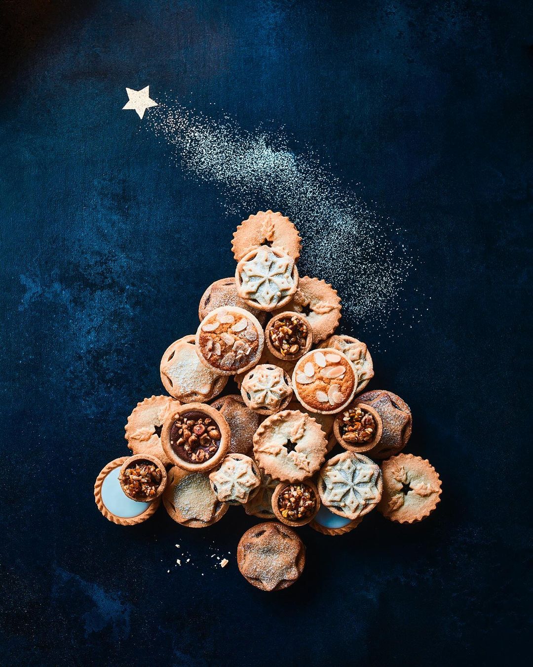 Overhead shot of a Christmas tree made of various shaped mince pies arranged on a dark blue surface with a star and glitter.