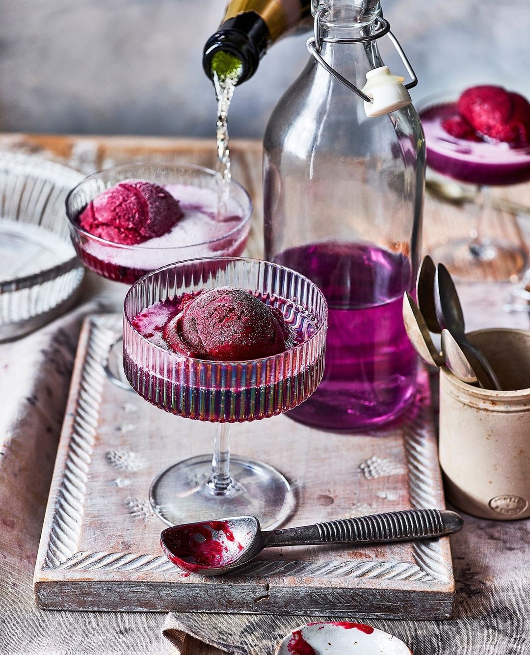 A glass bowl with red sorbet is on a rustic wooden tray, being poured with sparkling wine, surrounded by other glassware and silverware.