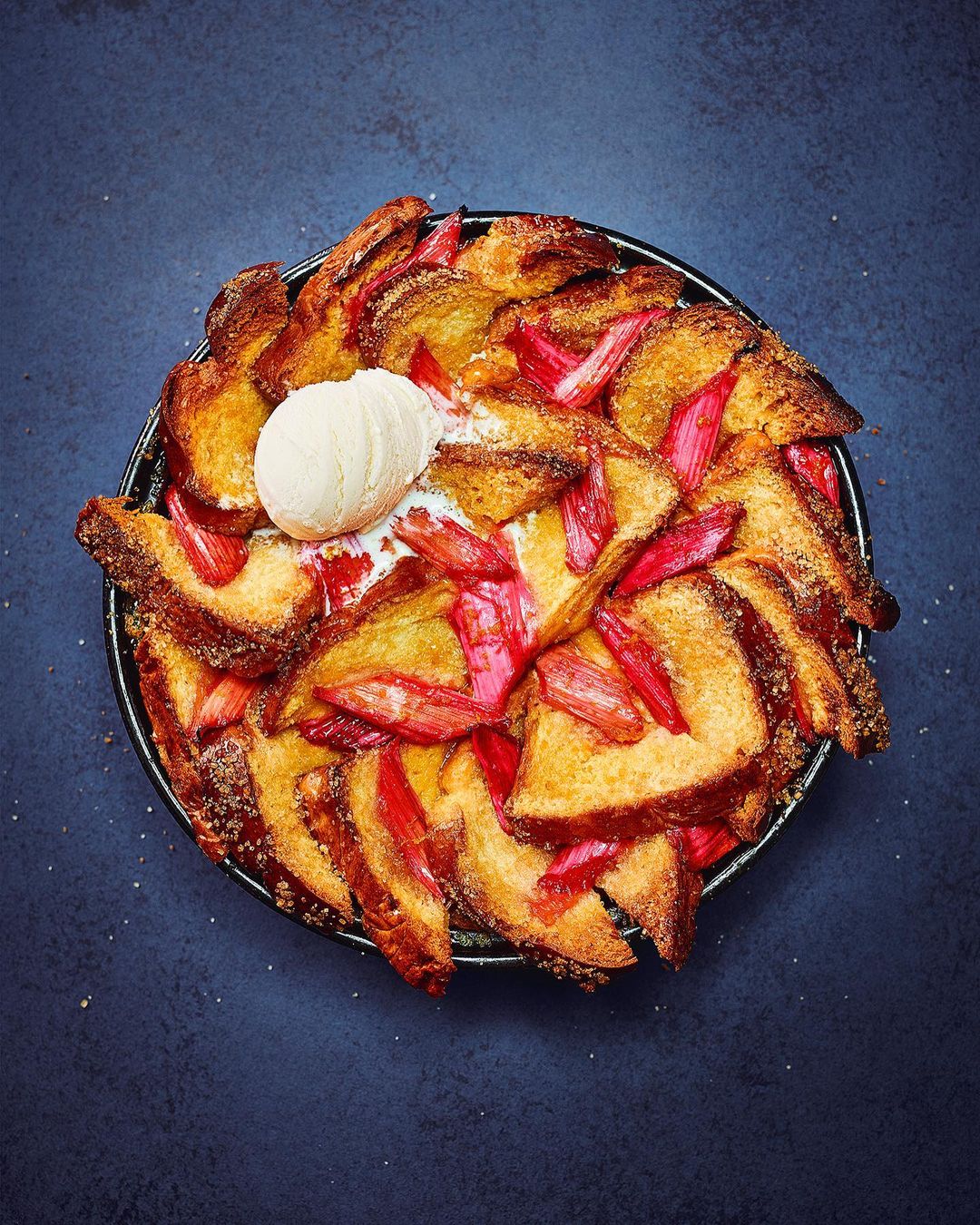 Overhead shot of a homemade rhubarb and custard bread pudding with a scoop of vanilla ice cream on top, placed on a dark background.