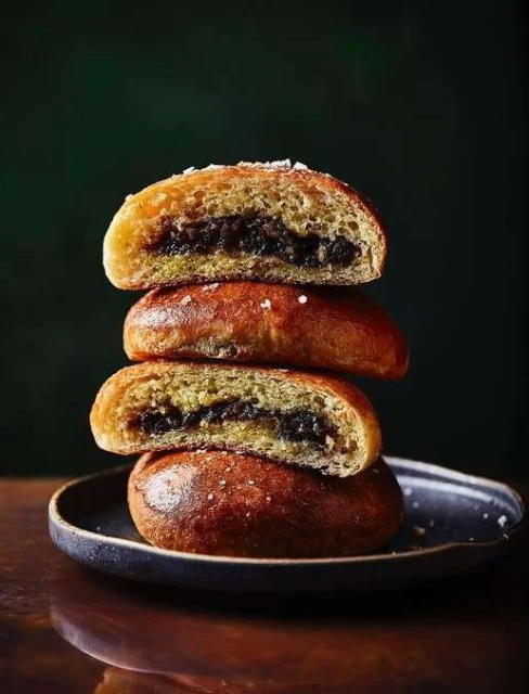 Tower of three enriched buns filled with a dark jam, stacked on a dark plate atop a reflective dark surface. High-contrast food shot.
