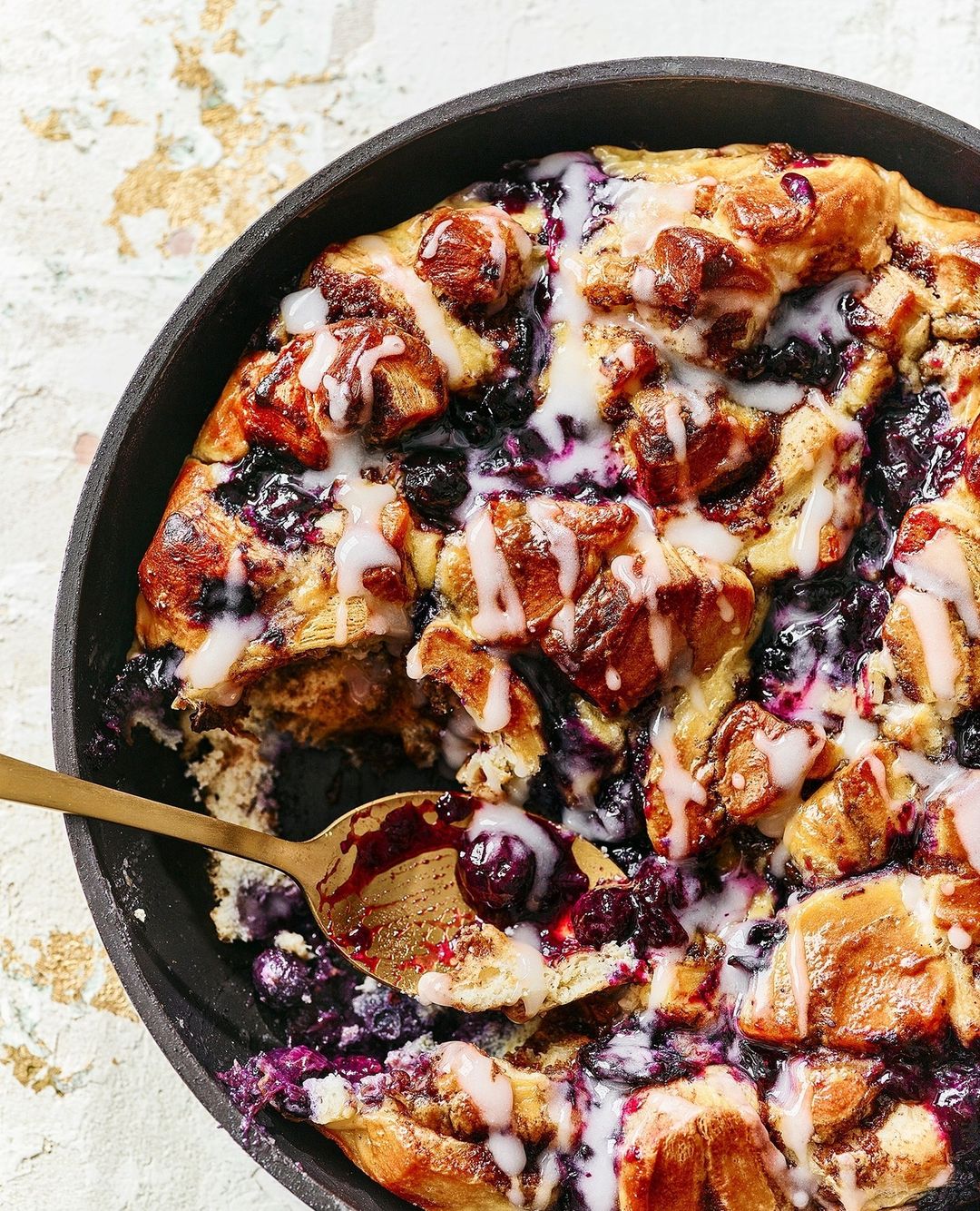 Overhead view of blueberry bread pudding in a cast iron skillet, topped with white icing drizzle and a golden spoon.