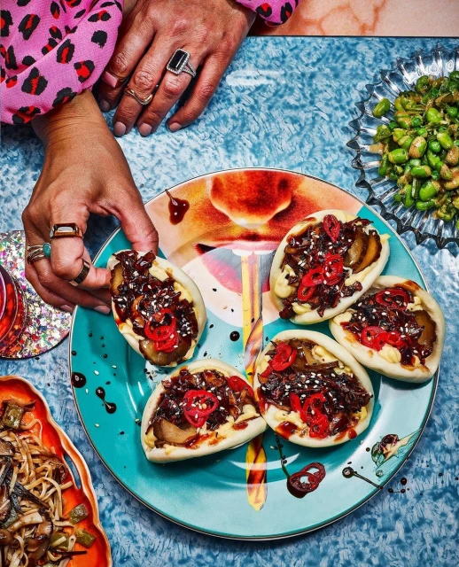 Overhead shot of a woman's hands holding a colorful plate of bao buns topped with meat and peppers surrounded by appetizer dishes.