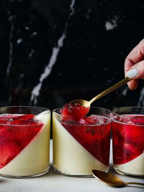 Three glasses of red and white panna cotta dessert are shown in a food photography studio setting, illuminated by natural light.