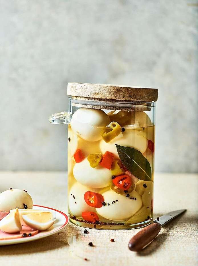 A glass jar filled with pickled eggs, red and yellow peppers, and spices, sits on a table with a plate of sliced eggs.